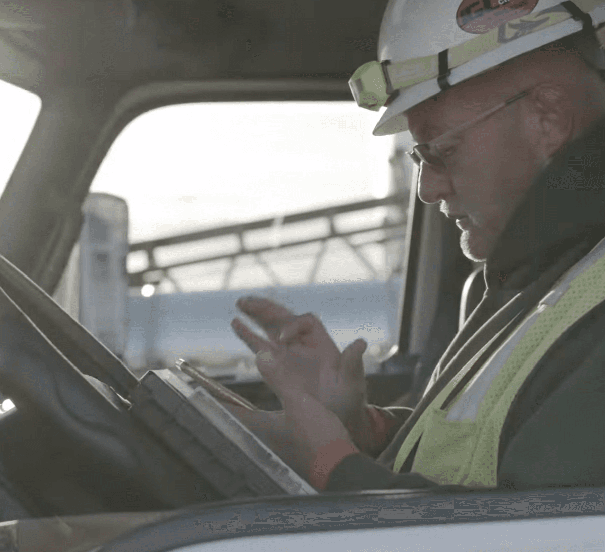 Driver in a truck cab using a tablet while wearing a hard hat and safety vest.